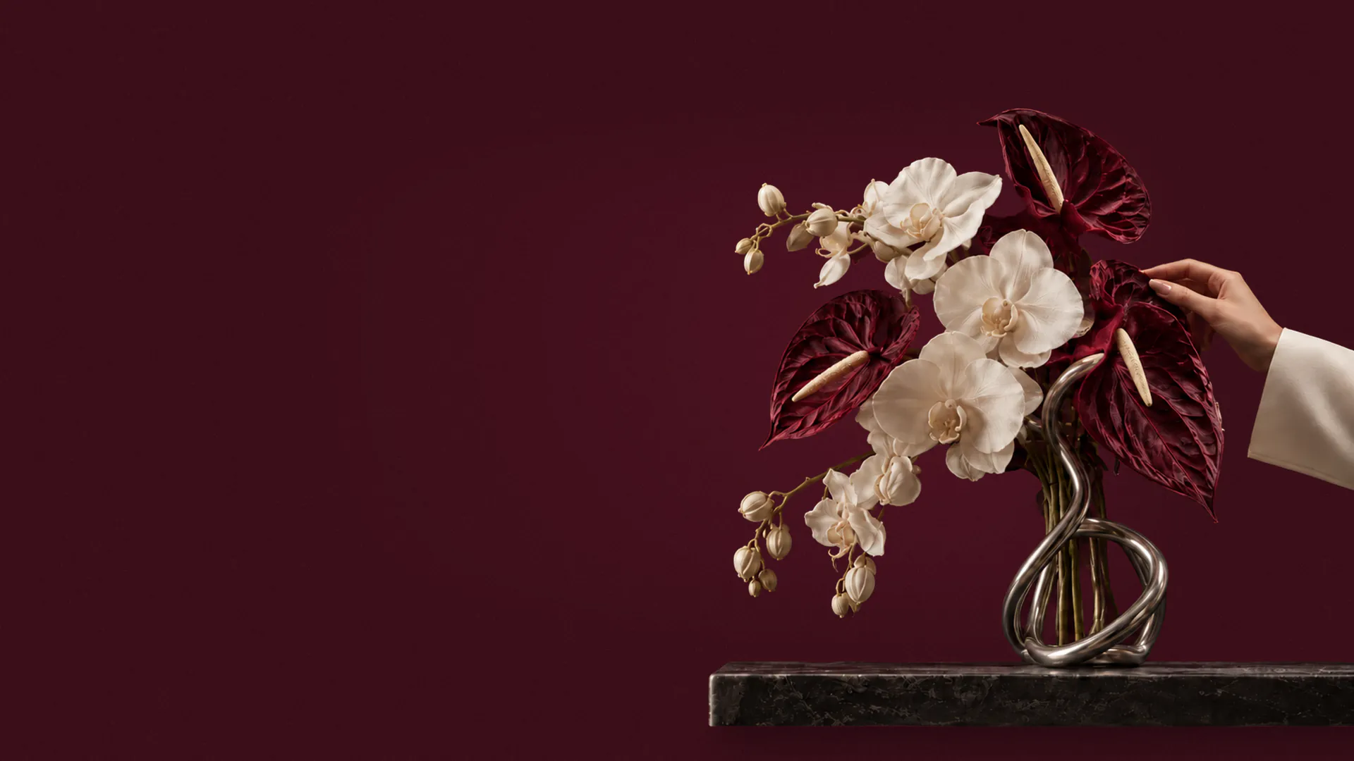Ivory silk orchids and burgundy faux anthurium in a chrome knot-shaped vase on a dark marble shelf, a hand in a white sleeve adjusting a leaf